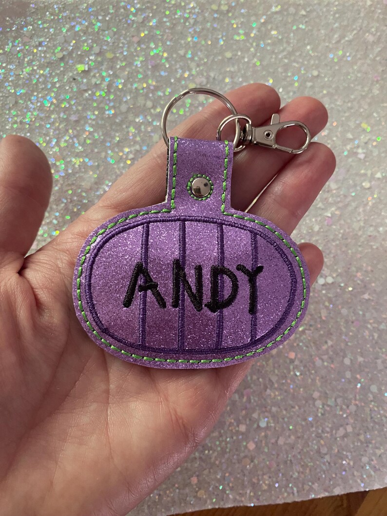 May include: A purple glittery oval keychain featuring vertical dark purple stripes and the name ANDY embroidered in black thread. The keychain is attached to a silver metal split ring and a lobster clasp, with bright green stitching along the edges.