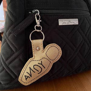 May include: A gold glittery keychain shaped like a bowling pin with the name ANDY embroidered in black thread. The keychain is attached to a silver metal lobster clasp and hangs from the front zipper of a black quilted fabric backpack.