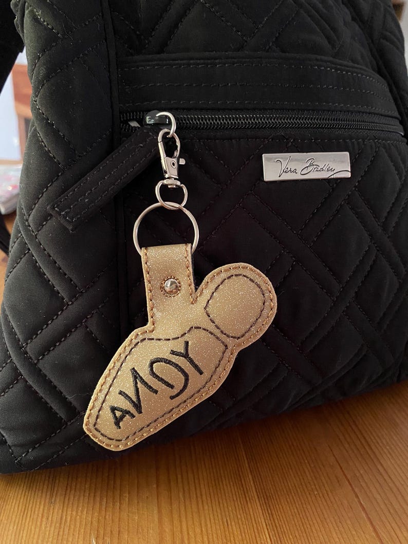 May include: A gold glittery keychain shaped like a bowling pin with the name ANDY embroidered in black thread. The keychain is attached to a silver metal lobster clasp and hangs from the front zipper of a black quilted fabric backpack.