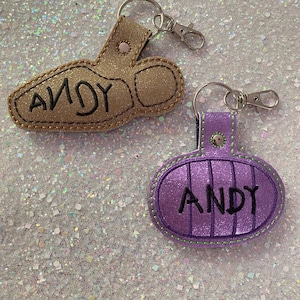 May include: Two glittery keychains with metal lobster clasps. One is a gold-colored boot shape with the name ANDY embroidered in black. The other is a purple oval shape with vertical stitching and the name ANDY embroidered in black.