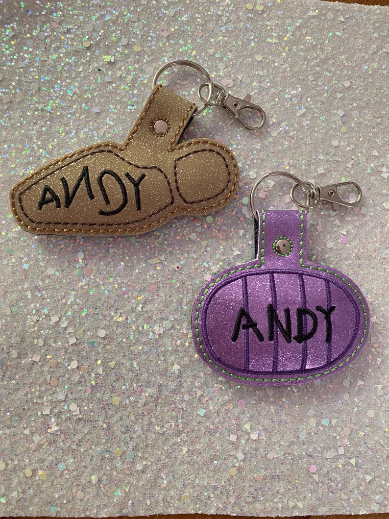 May include: Two glittery keychains with metal lobster clasps. One is a gold-colored boot shape with the name ANDY embroidered in black. The other is a purple oval shape with vertical stitching and the name ANDY embroidered in black.
