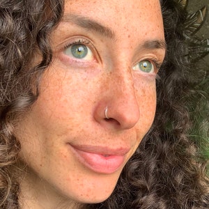 Può includere: Una persona con capelli castani ricci e occhi verdi, che indossa una felpa nera. La persona ha un piercing al naso e lentiggini. Una pianta verde è visibile sullo sfondo.