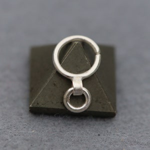 May include: A silver-colored metal charm with a small ring attached to a larger ring, connected by a short bar. The charm is set on a dark green, pyramid-shaped object. The background is a solid gray.