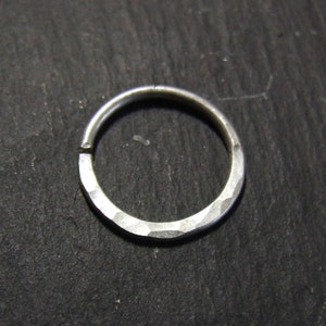 Peut inclure: Une bague en argent avec une texture martelée. La bague est circulaire avec un petit espace et repose sur une surface sombre et texturée. La surface de la bague a un aspect facetté, captant la lumière.