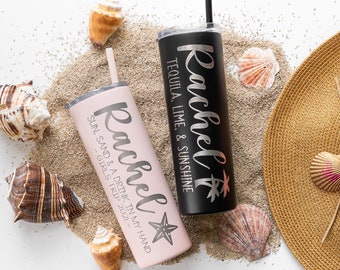 May include: Two personalized stainless steel tumblers with the name "Rachel" on them. One tumbler is black with the text "Tequila, Lime, & Sunshine" and the other is pink with the text "Sun, Sand & A Drink In My Hand - Girls Trip 2021 - ". The tumblers are sitting on a bed of sand with seashells.
