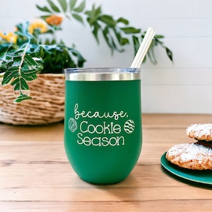 May include: Green stainless steel tumbler with a white inscription that reads "because, Cookie Season". The tumbler has a straw and is sitting on a wooden surface with a plate of cookies.