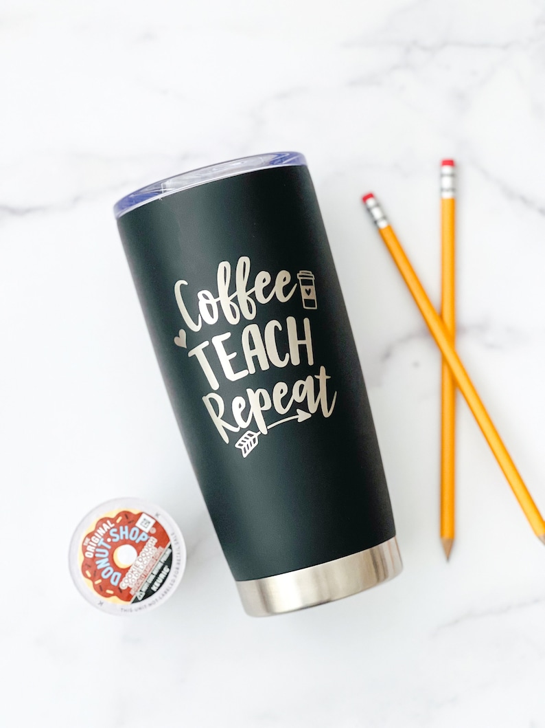 K&ouml;nnte beinhalten: Eine schwarze Edelstahl-Reise-Thermoskanne mit silbernem Deckel und einer goldenen Aufschrift "Coffee Teach Repeat" mit einem kleinen Kaffeetassen-Symbol und einem Pfeil.