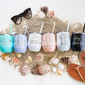K&ouml;nnte beinhalten: Sechs Edelstahlbecher mit verschiedenen sommerlichen Designs und Spr&uuml;chen. Die Becher sind in einer Reihe auf einem Sandbett mit verstreuten Muscheln angeordnet. Die Becher zeigen Spr&uuml;che wie "Sippin' on Sunshine", "Summer Vibes", "Life is Better at the Pool" und "Day Drinkin".