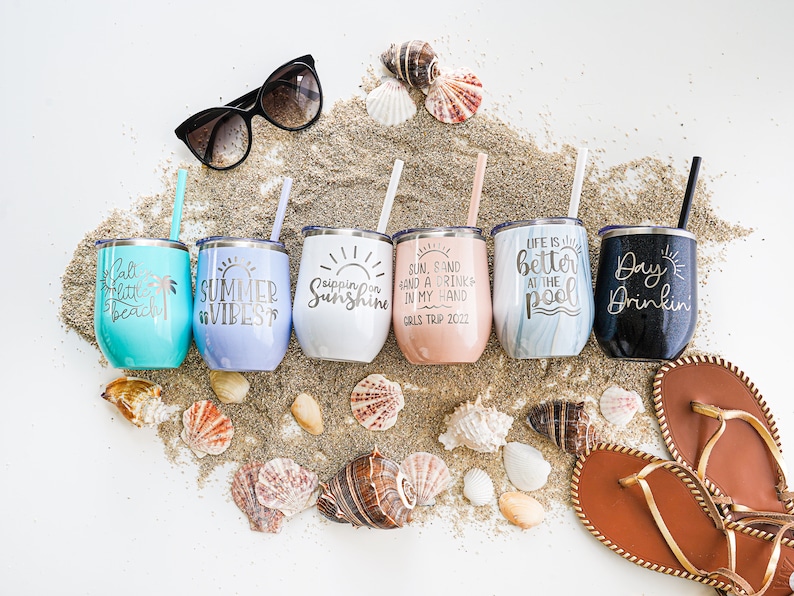 K&ouml;nnte beinhalten: Sechs Edelstahlbecher mit verschiedenen sommerlichen Designs und Spr&uuml;chen. Die Becher sind in einer Reihe auf einem Sandbett mit verstreuten Muscheln angeordnet. Die Becher zeigen Spr&uuml;che wie "Sippin' on Sunshine", "Summer Vibes", "Life is Better at the Pool" und "Day Drinkin".