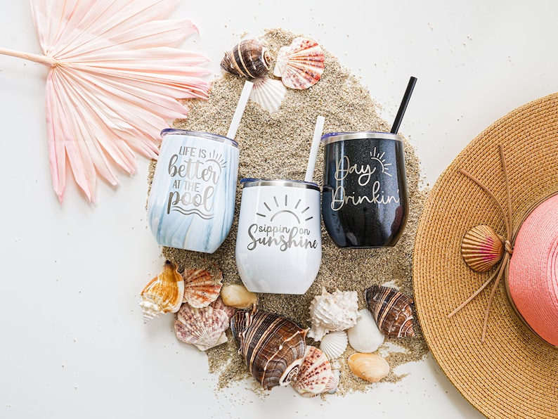 K&ouml;nnte beinhalten: Drei Edelstahlbecher mit sommerlichen Spr&uuml;chen. Die Becher stehen auf einem Sandbett mit Muscheln und einem Strohhut. Die Becher sagen "Life is better at the pool", "Sipping on Sunshine" und "Day Drinkin".