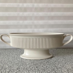 May include: Cream-coloured, pedestal serving bowl with two handles. The bowl has a ribbed design around the sides and a wide, oval shape. The pedestal base is round and supports the bowl. The background is a neutral, striped pattern.