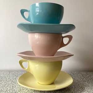May include: Four pastel-coloured teacups and saucers stacked. The colours are light blue, pink, and yellow. Each cup has a curved handle. The saucers are square with rounded edges. The cups and saucers are stacked on top of each other.