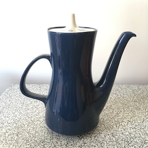 May include: A navy blue ceramic coffee pot with a curved handle and a long spout. The lid is off-white with a floral design. The pot is on a patterned surface, evoking a vintage or retro style. The pot is likely around 25 cm tall.