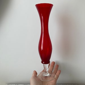 May include: A tall, elegant red glass vase with a flared top and a clear, textured base. The vase has a slender, hourglass shape and stands approximately 30 cm tall. The base is clear glass with a decorative, textured design.