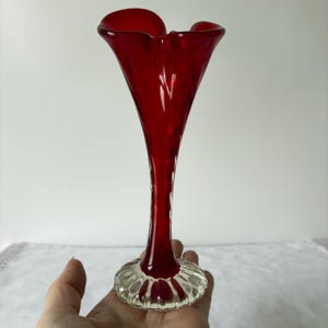 May include: A tall, slender glass vase featuring a deep red, trumpet-shaped top that transitions into a clear, ribbed, circular base. The glass has a smooth, polished finish, with the vibrant red colour concentrated in the upper section and neck.