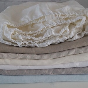 Peut inclure: Une pile de linge de lit en lin dans différentes nuances de blanc, beige et bleu clair. La couche supérieure est un drap housse blanc avec un bord festonné.