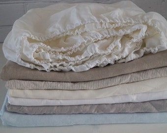 May include: A stack of linen bedding in various shades of white, beige, and light blue. The top layer is a white fitted sheet with a ruffled edge.