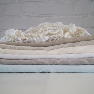 May include: A stack of folded linen fabric in various shades of white, beige, and light blue. The fabric is soft and textured, with a subtle sheen.