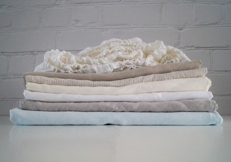 May include: A stack of folded linen fabric in various shades of white, beige, and light blue. The fabric is soft and textured, with a subtle sheen.