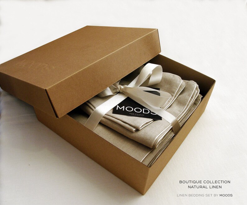 May include: A brown cardboard box with a white ribbon and a tag that says "MOODS" is open to reveal a set of natural linen bedding. The bedding is folded neatly inside the box. The box is labelled "Boutique Collection Natural Linen Linen Bedding Set by MOODS".