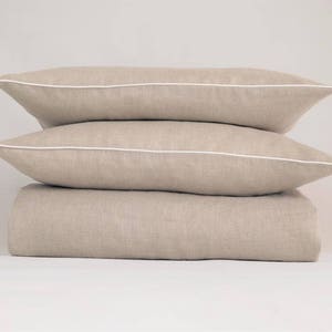 May include: A stack of three beige linen pillowcases with white piping. The pillowcases are arranged in a pyramid shape.