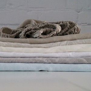 May include: A stack of six linen sheets in various shades of beige, white, and light blue. The top sheet is folded and has a gathered edge.