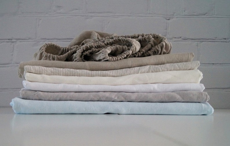 May include: A stack of six linen sheets in various shades of beige, white, and light blue. The top sheet is folded and has a gathered edge.