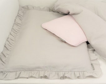 May include: A gray linen crib sheet with a ruffled edge and a pink pillow. The sheet has a small gray tag with the word "Crib" on it.