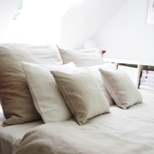 May include: A white bed with a white linen duvet cover and several white and beige pillows. The bed is against a white wall and there is a wooden bedside table next to the bed.