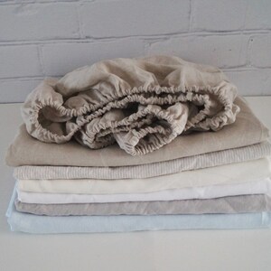 May include: A stack of linen bed sheets in various shades of beige, white, and light blue. The top sheet is folded and has a gathered edge.