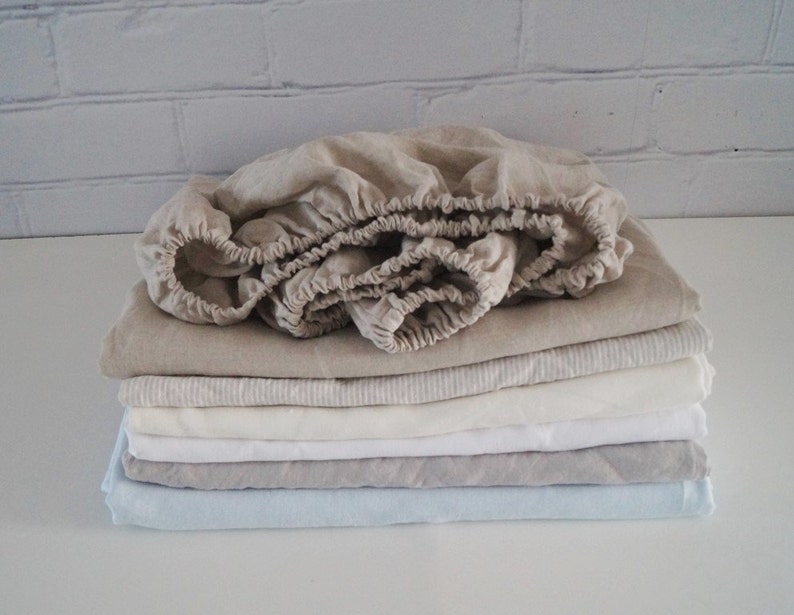 May include: A stack of linen bed sheets in various shades of beige, white, and light blue. The top sheet is folded and has a gathered edge.