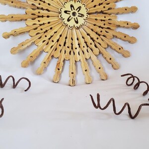 May include: A sun-shaped wall decor made from wooden clothespins with a small wooden flower in the center. The decor is on a white background.