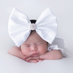 May include: A large white satin bow headband featuring a circular center embellishment covered in small clear crystals. The bow is structured with wide, stiffened fabric loops that stand upright, creating a prominent decorative accessory.