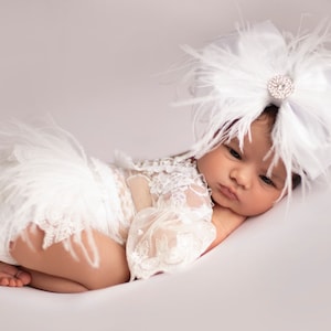 May include: A newborn baby wearing a white lace romper with feather accents and a large white bow headband adorned with a sparkling jewel. The baby is lying on a white surface.