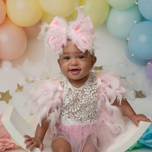 May include: A baby wearing a pink feather-trimmed outfit with a white lace bodice and a tulle skirt. The outfit includes a large pink bow headband with a rhinestone accent. Pastel balloons and gold stars decorate the background.