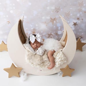 May include: A newborn baby sleeps peacefully in a cream-colored crescent moon prop. The baby wears a white lace outfit and a large white bow. The moon is surrounded by gold stars and fluffy white clouds, creating a dreamy scene.