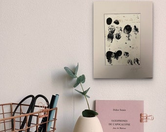 May include: A copper-colored wire basket holds black scissors and a blue pencil. A white, oval vase with a sprig of greenery sits next to a pink book titled "SAXOPHONES DE L'APOCALYPSE". A framed black and white abstract print hangs on the wall.