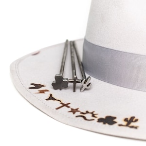 Puede incluir: Un sombrero de vaquero de fieltro blanco con una banda gris y un juego de cuatro hierros de marcar de metal con diferentes diseños, incluyendo un trébol, una cruz, un cactus y una herradura.