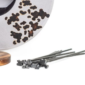 Op de afbeelding: Een set van 10 metalen gereedschappen met een uniek ontwerp, dat lijkt op een gestileerde vogel, is gerangschikt op een wit oppervlak. De gereedschappen zijn gemaakt van donker metaal en hebben een glanzende afwerking. Een witte hoed met een zwart en bruin patroon is zichtbaar op de achtergrond.