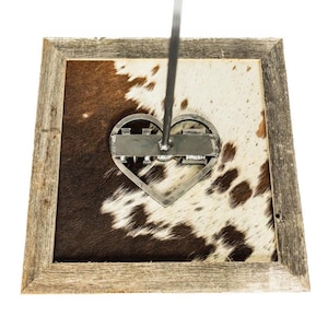 May include: A rustic wooden framed piece featuring a cowhide pattern in shades of brown and white. A metal heart-shaped brand is centered on the hide. The frame has a weathered appearance, adding to the piece's country aesthetic.