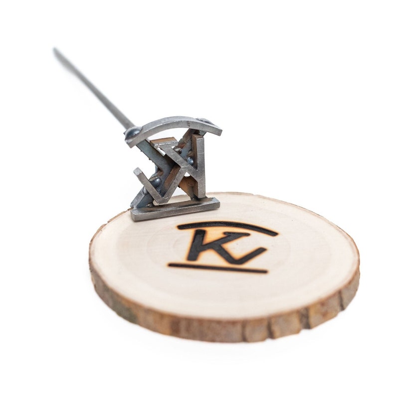 May include: A metal branding iron with a letter K design, used to create a custom wood burn on a wooden disc.