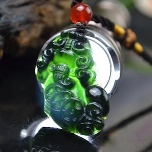 May include: A green jade pendant carved in the shape of a mythical creature. The pendant is on a black cord with a red bead.