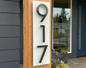 Puede incluir: Un letrero rectangular blanco con los números 9, 1 y 7 en negro. Los números están en una fuente sans-serif moderna. El letrero está montado en un poste de madera, en el exterior, cerca de una puerta principal.