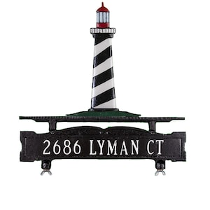 May include: Black metal mailbox with a lighthouse design. The mailbox has a black and white striped lighthouse with a red roof and a green base. The mailbox also has a black metal sign that says "2686 LYMAN CT".