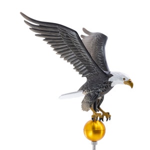 May include: A bald eagle sculpture with dark gray wings, a white head, and a yellow beak, perched atop a smooth, spherical gold-colored finial. The eagle is depicted with its wings fully extended in an upward position, featuring detailed feather textures.