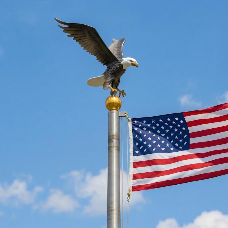 May include: A metal bald eagle statue with outstretched wings perched atop a gold-colored sphere on a silver flagpole. The American flag with its red, white, and blue stripes and white stars on a blue field is attached to the pole below the eagle.