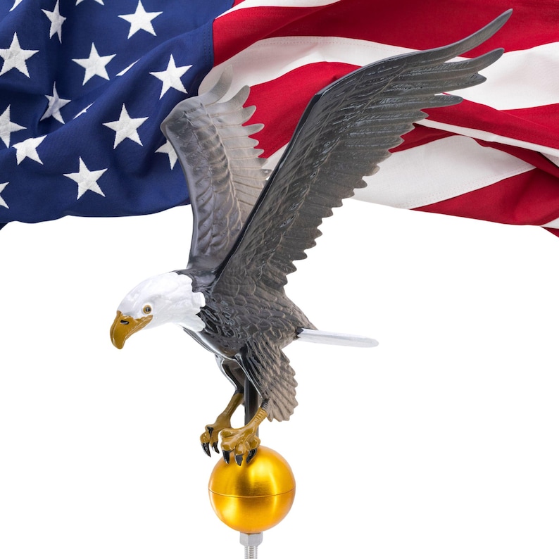 May include: A three-dimensional bald eagle figure with a white head, yellow beak, and dark brown wings spread wide in flight. The eagle is perched atop a smooth, spherical gold-colored finial, positioned in front of a folded American flag.