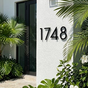 May include: Black metal house numbers, 1748, mounted on a textured white wall. The numbers are in a modern font. The wall is next to a black door and windows, with lush green plants and white flowers in the foreground.