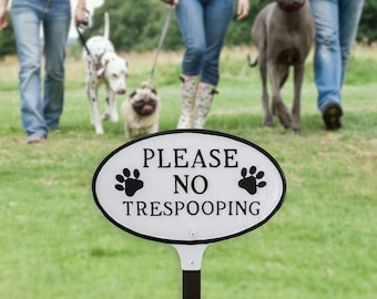 May include: White oval sign with black paw prints and the text "PLEASE NO TRESPOOPING". The sign is on a black metal pole and is in a grassy area with people and dogs walking in the background.