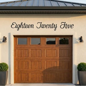 May include: A wooden garage door with a brown finish and rectangular panels. Above the door, the words "Eighteen Twenty Five" are displayed in a black, cursive font. The garage is framed by a light-colored wall and a dark roof.